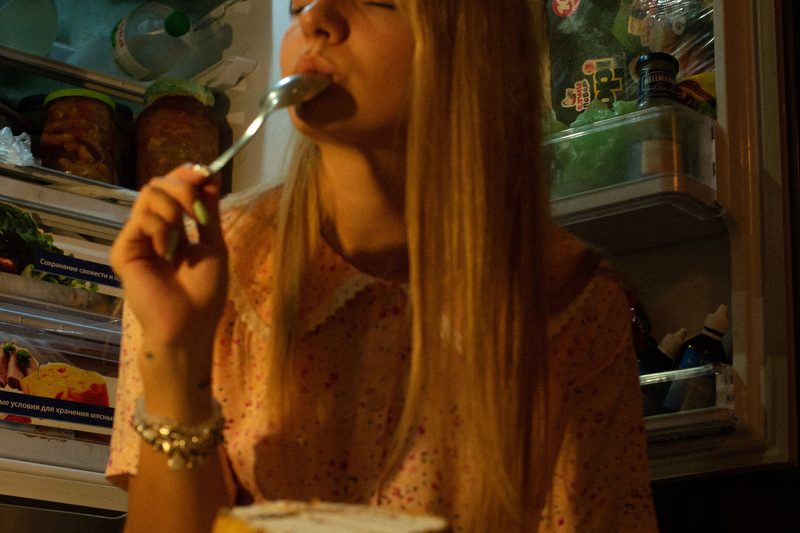 Femme dégustant une part de gâteau devant un frigo ouvert la nuit