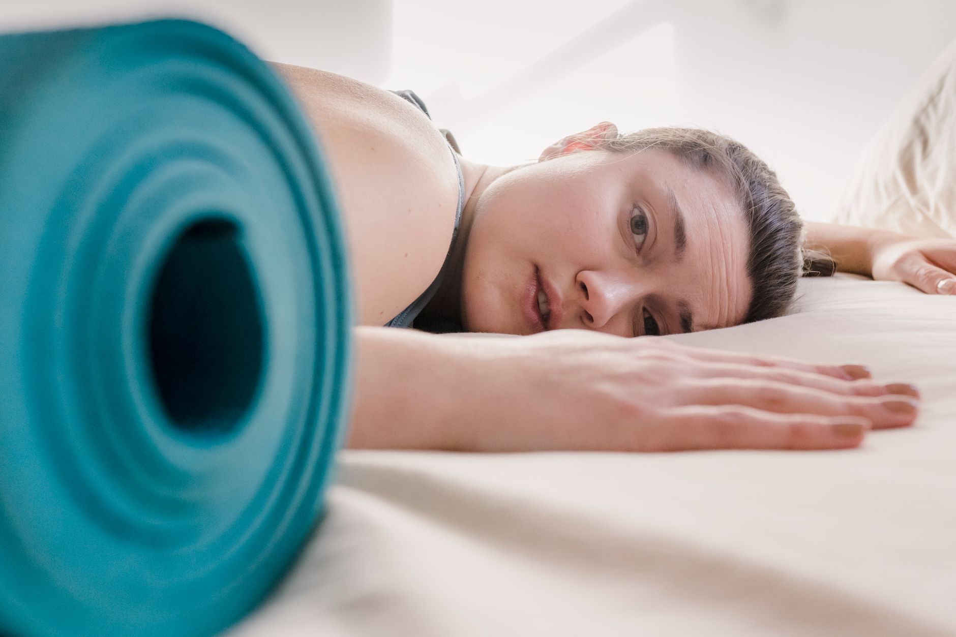 Femme allongée au sol près d'un tapis de sport enroulé, le regard fatigué, incapable de commencer sa séance