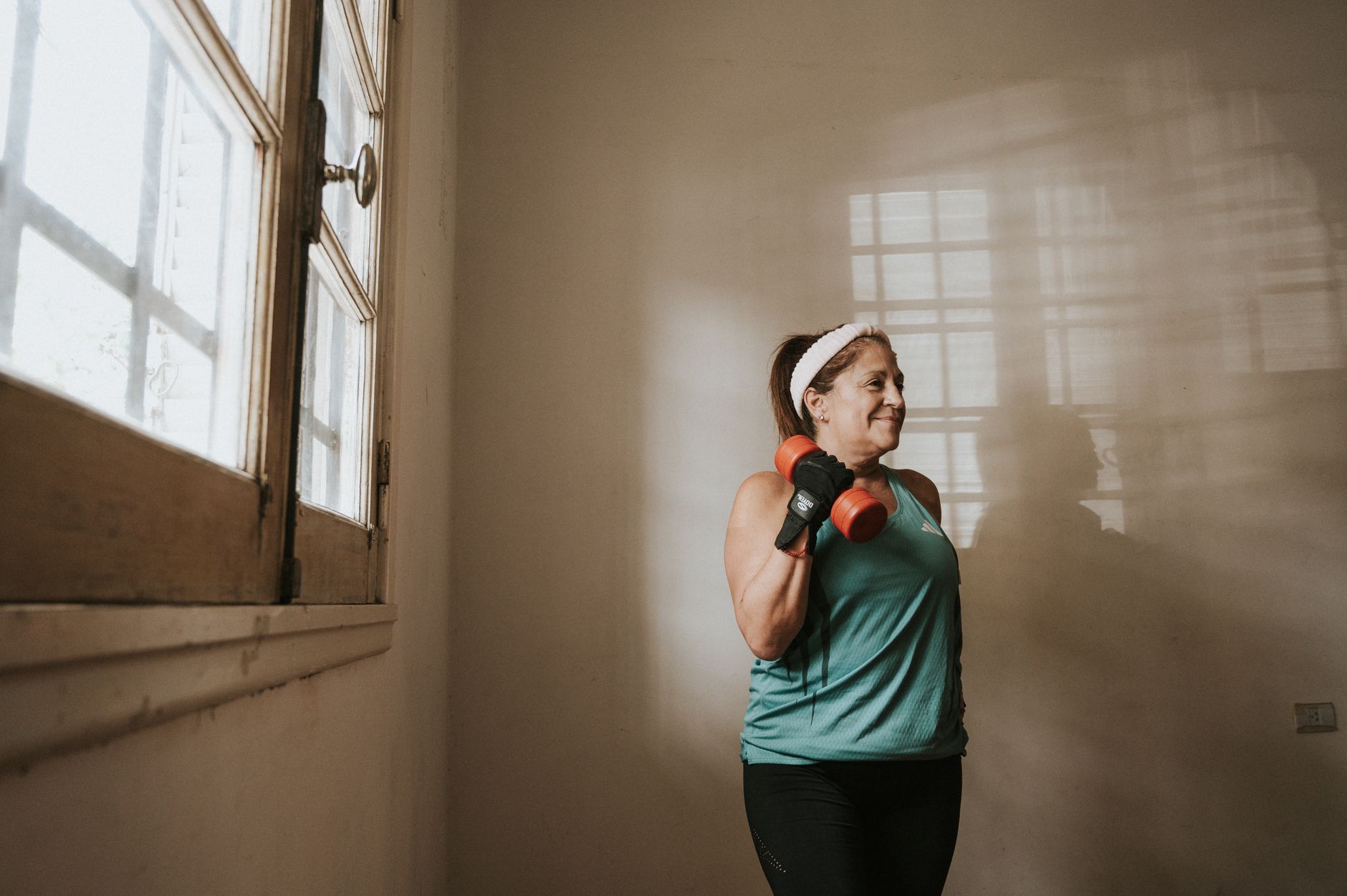Femme d'une quarantaine d'années souriante, faisant un exercice de renforcement avec des haltères chez elle