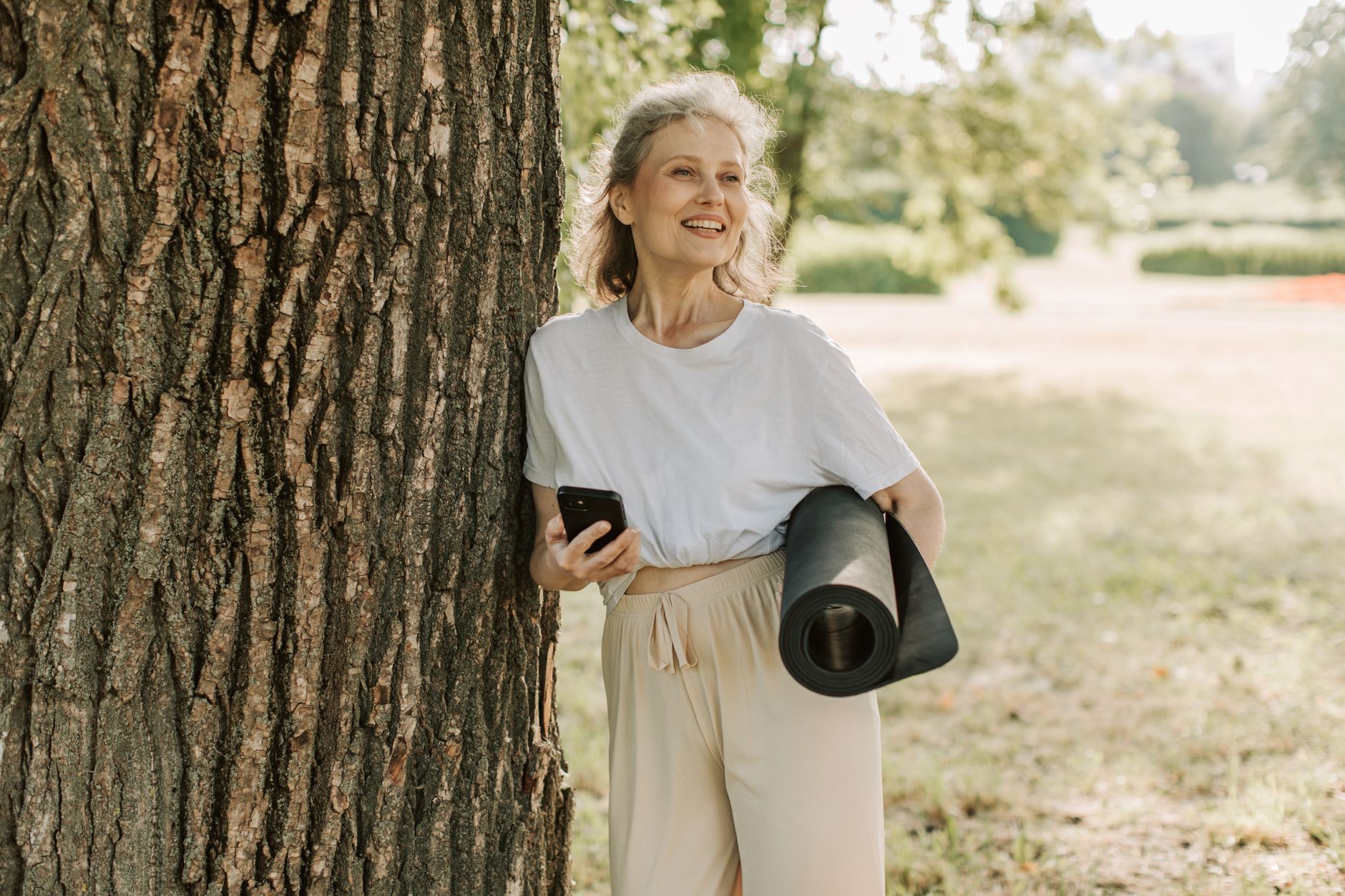 Femme souriante et active, appuyée contre un arbre dans un parc, tapis de yoga sous le bras
