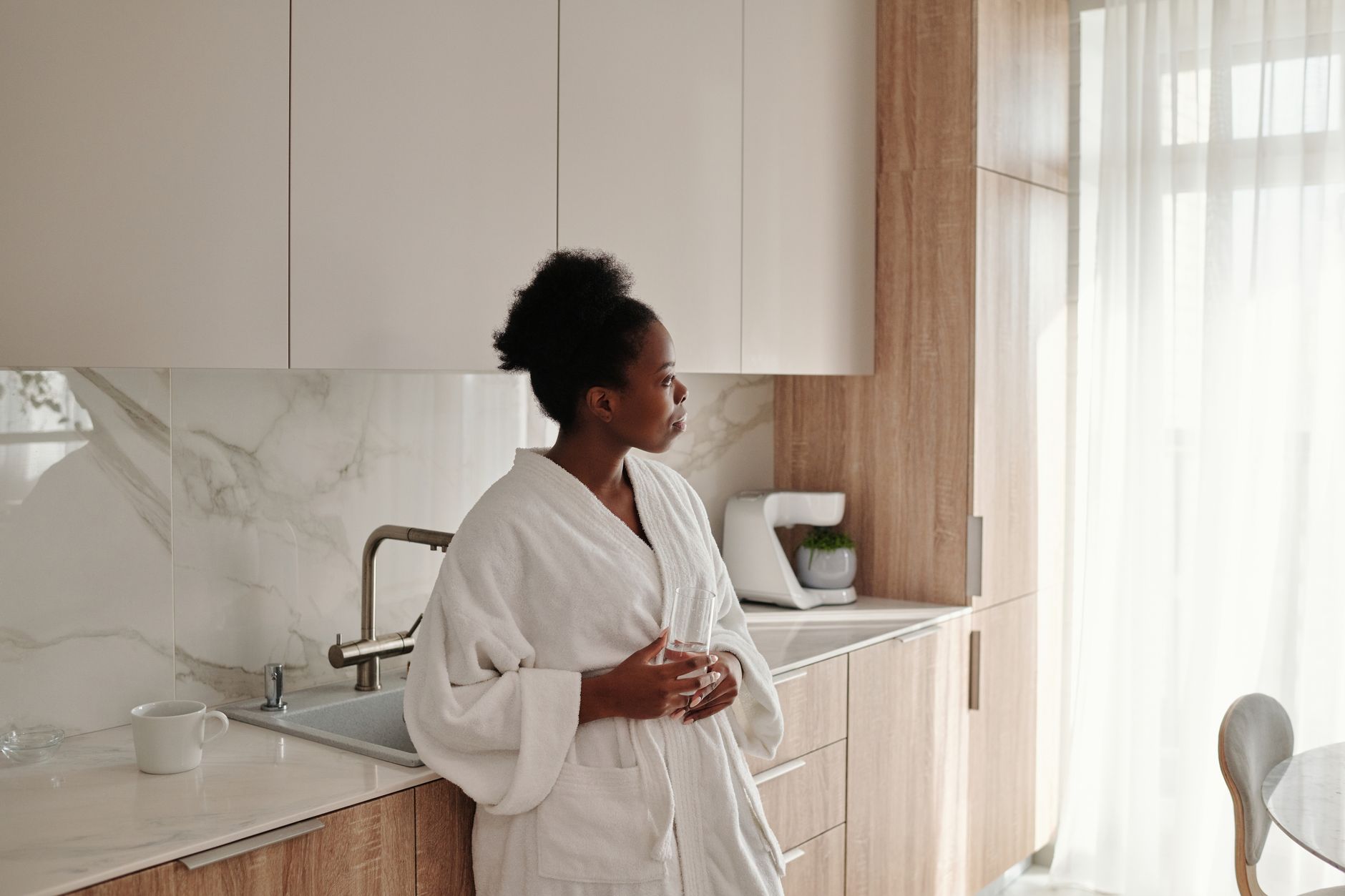 Femme en peignoir blanc dans une cuisine lumineuse, debout près de la fenêtre avec une tasse à la main, moment calme du matin